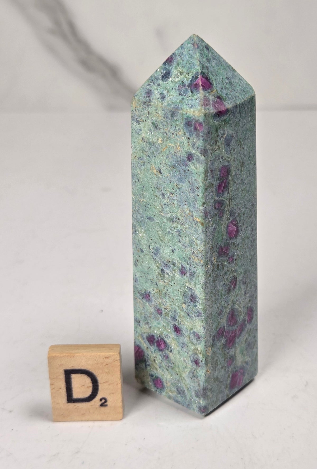 Green crystal polished point with pink and purple spots next to a scrabble tile against a white background