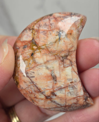 Hand holding a polished crystal moon carving with tan, yellow and red coloring against a white background