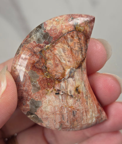Hand holding a polished crystal moon carving with tan, yellow and red coloring against a white background