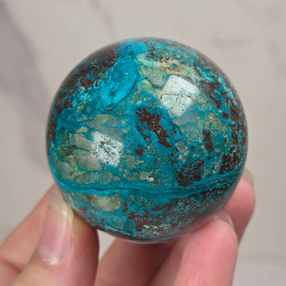 Hand holding a blue and brown crystal polished sphere carving against a white background