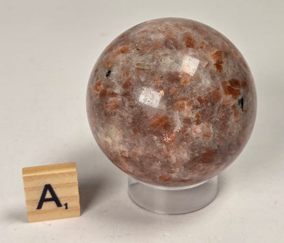 Orange colored crystal sphere carving on an acrylic stand with a scrabble tile against a white background