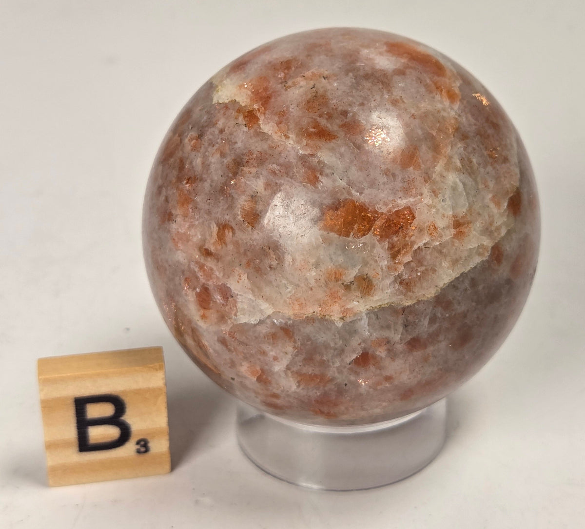 Orange colored crystal sphere carving on an acrylic stand with a scrabble tile against a white background
