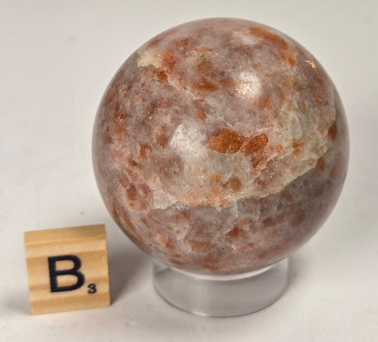 Orange colored crystal sphere carving on an acrylic stand with a scrabble tile against a white background