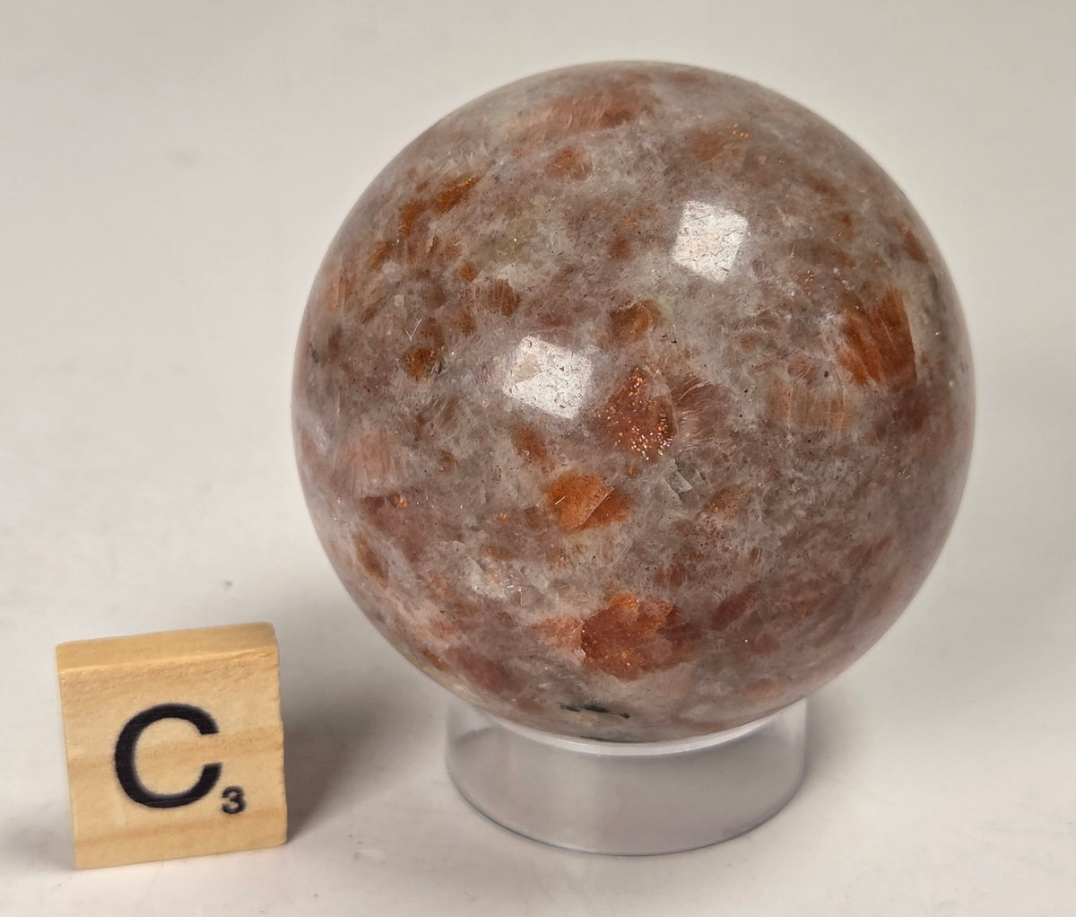 Orange colored crystal sphere carving on an acrylic stand with a scrabble tile against a white background