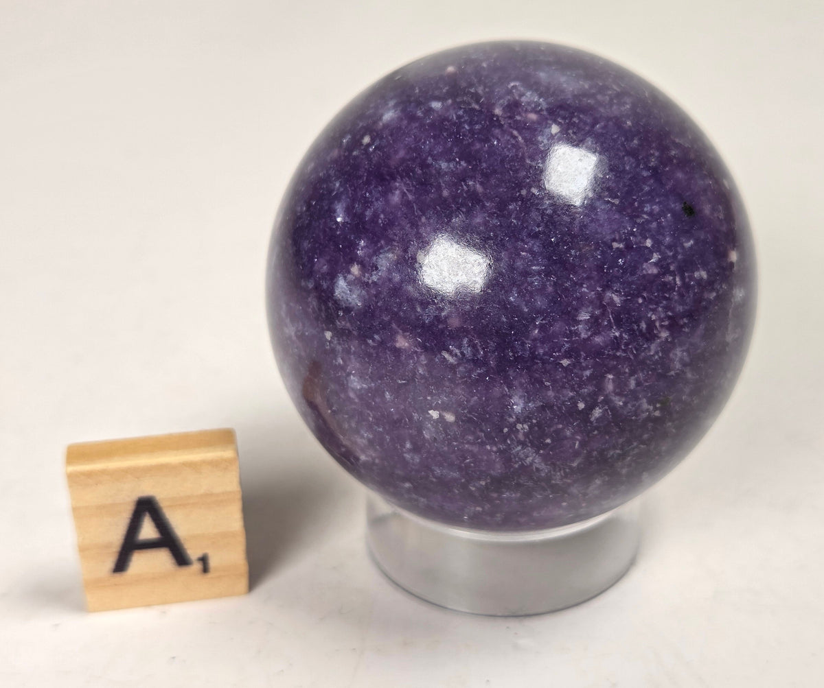 Purple crystal sphere carving on an acrylic stand with a scrabble tile against a white background