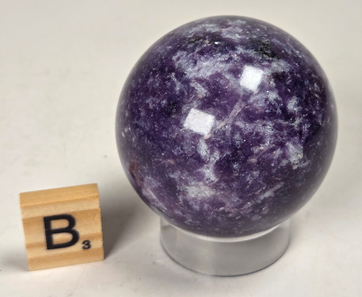 Purple crystal sphere carving on an acrylic stand with a scrabble tile against a white background