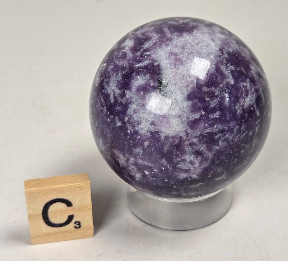 Purple crystal sphere carving on an acrylic stand with a scrabble tile against a white background