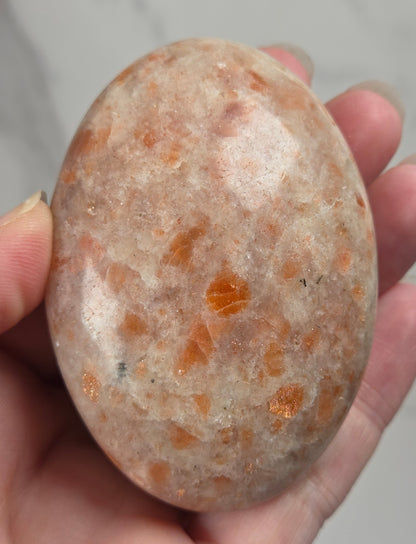 Polished orange sunstone free form carving being held in a hand against a marbled background