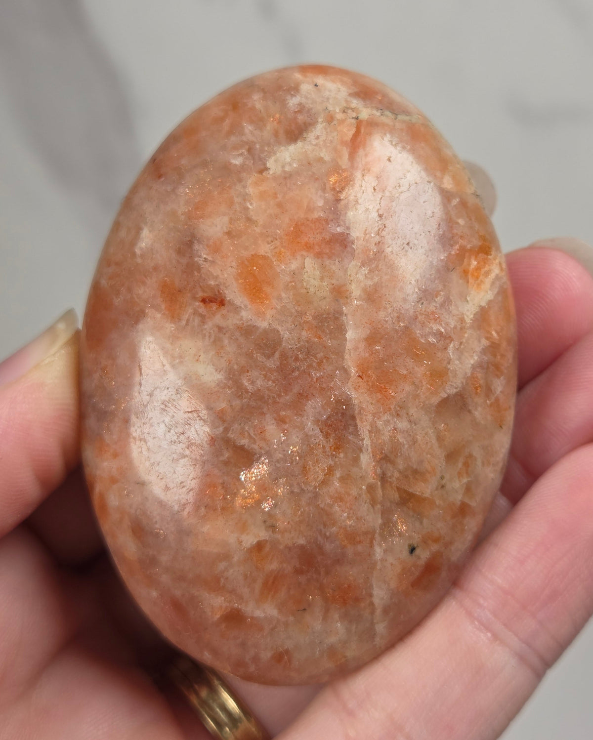 Polished orange sunstone free form carving being held in a hand against a marbled background