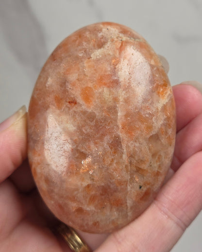 Polished orange sunstone free form carving being held in a hand against a marbled background