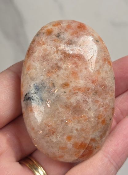 Polished orange sunstone free form carving being held in a hand against a marbled background