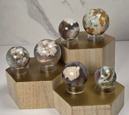 Collection of crystal sphere carvings with pink and grey coloring, each sphere on an acrylic holder, sitting on a wooden stand against a marbled background.