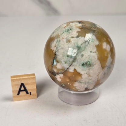 Flower Agate crystal sphere carving with pink and grey coloring, on an acrylic holder, with a scrabble tile against a marbled background.