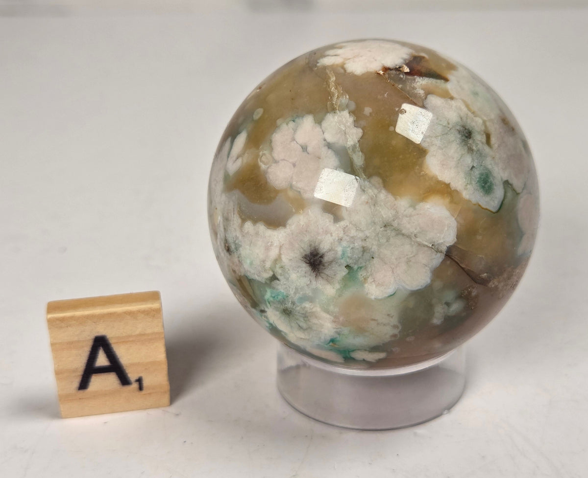 Flower Agate crystal sphere carving with pink and grey coloring, on an acrylic holder, with a scrabble tile against a marbled background.
