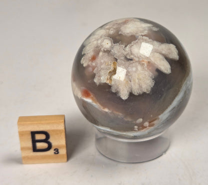 Flower Agate crystal sphere carving with pink and grey coloring, on an acrylic holder, with a scrabble tile against a marbled background.