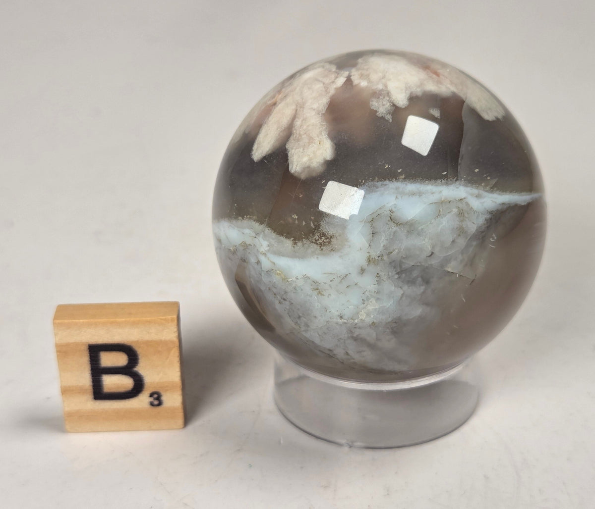 Flower Agate crystal sphere carving with pink and grey coloring, on an acrylic holder, with a scrabble tile against a marbled background.