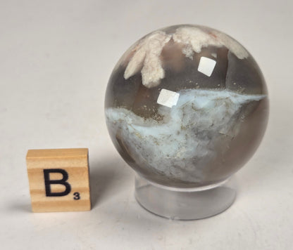 Flower Agate crystal sphere carving with pink and grey coloring, on an acrylic holder, with a scrabble tile against a marbled background.