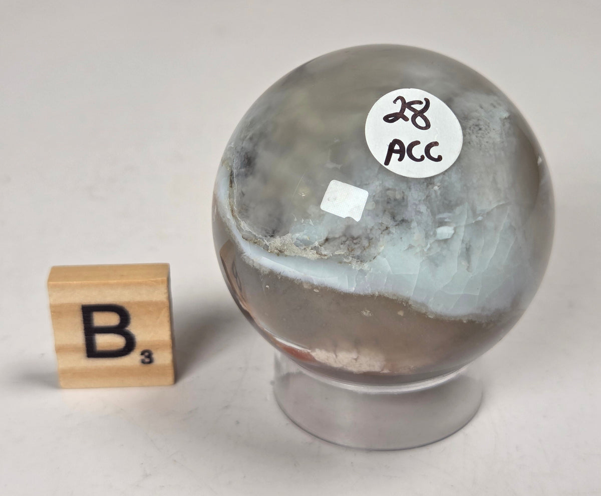 Flower Agate crystal sphere carving with pink and grey coloring, on an acrylic holder, with a scrabble tile against a marbled background.