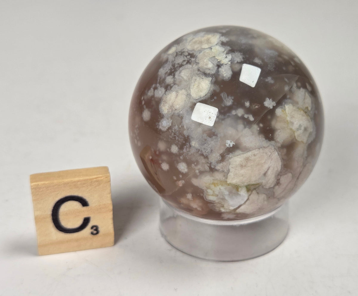 Flower Agate crystal sphere carving with pink and grey coloring, on an acrylic holder, with a scrabble tile against a marbled background.
