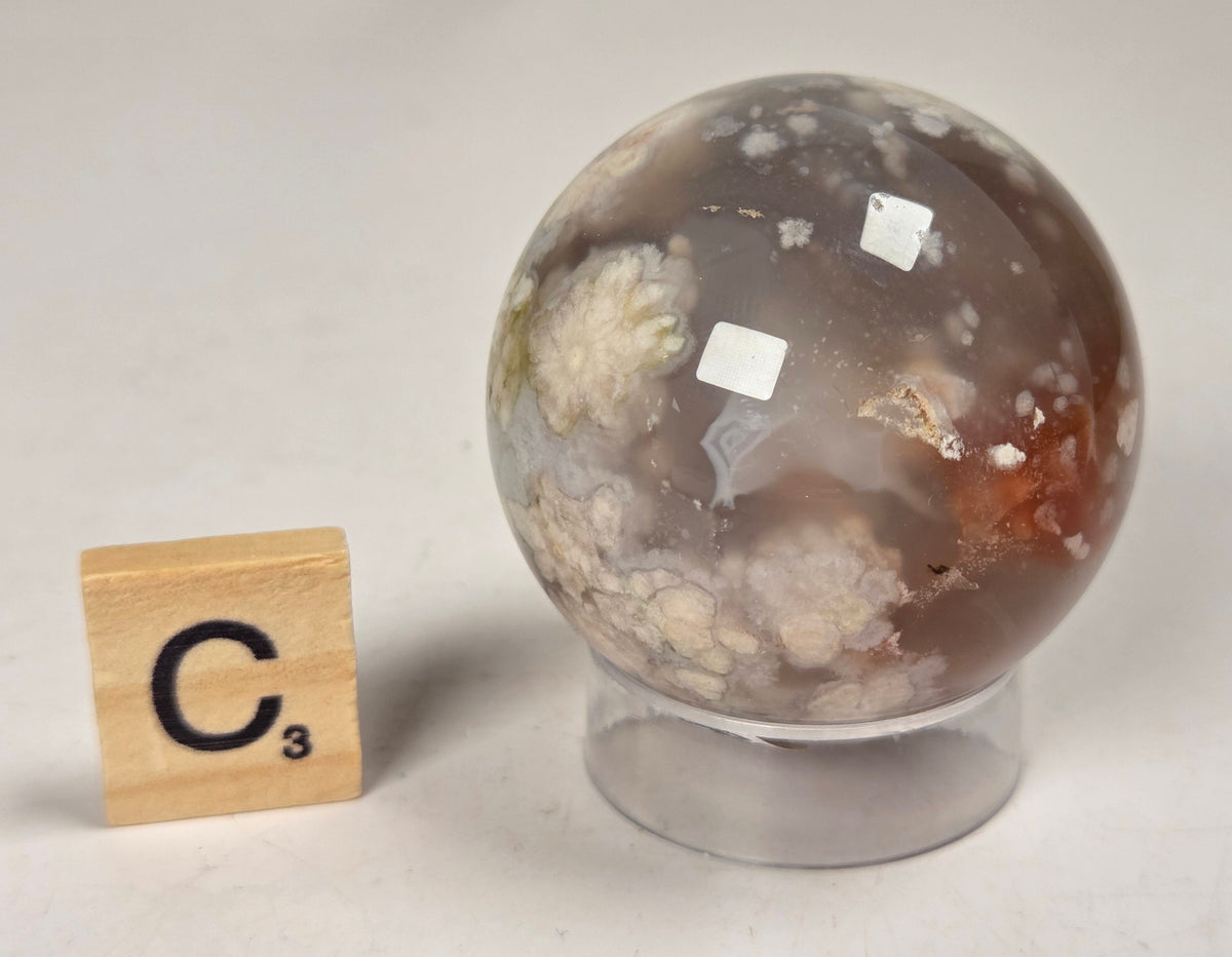 Flower Agate crystal sphere carving with pink and grey coloring, on an acrylic holder, with a scrabble tile against a marbled background.
