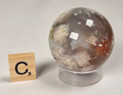 Flower Agate crystal sphere carving with pink and grey coloring, on an acrylic holder, with a scrabble tile against a marbled background.