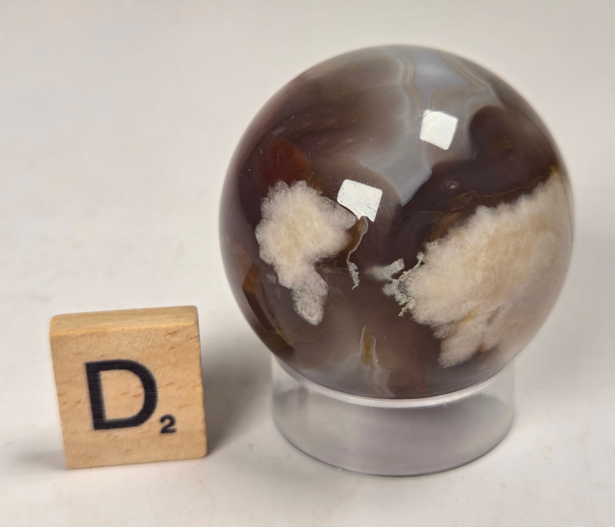 Flower Agate crystal sphere carving with pink and grey coloring, on an acrylic holder, with a scrabble tile against a marbled background.