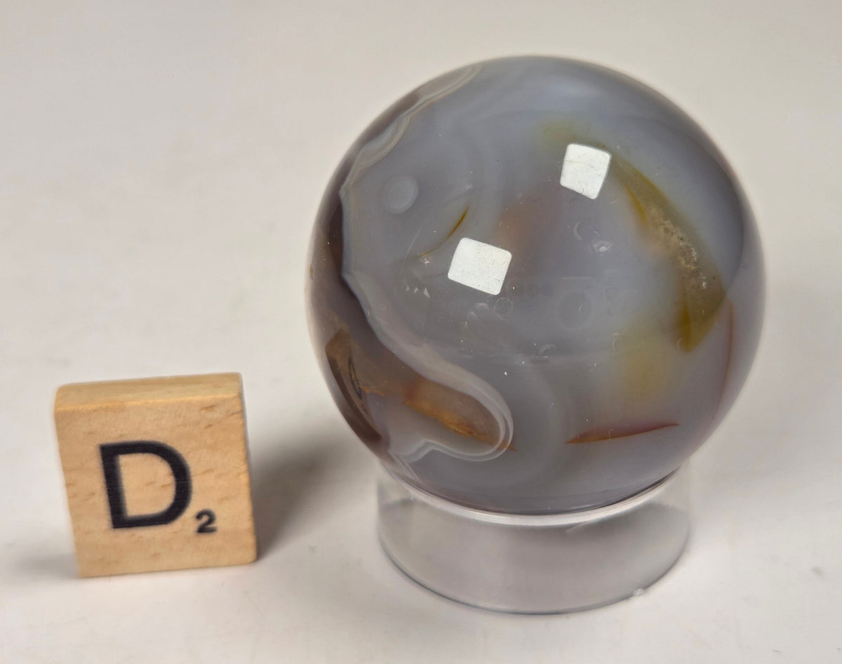 Flower Agate crystal sphere carving with pink and grey coloring, on an acrylic holder, with a scrabble tile against a marbled background.