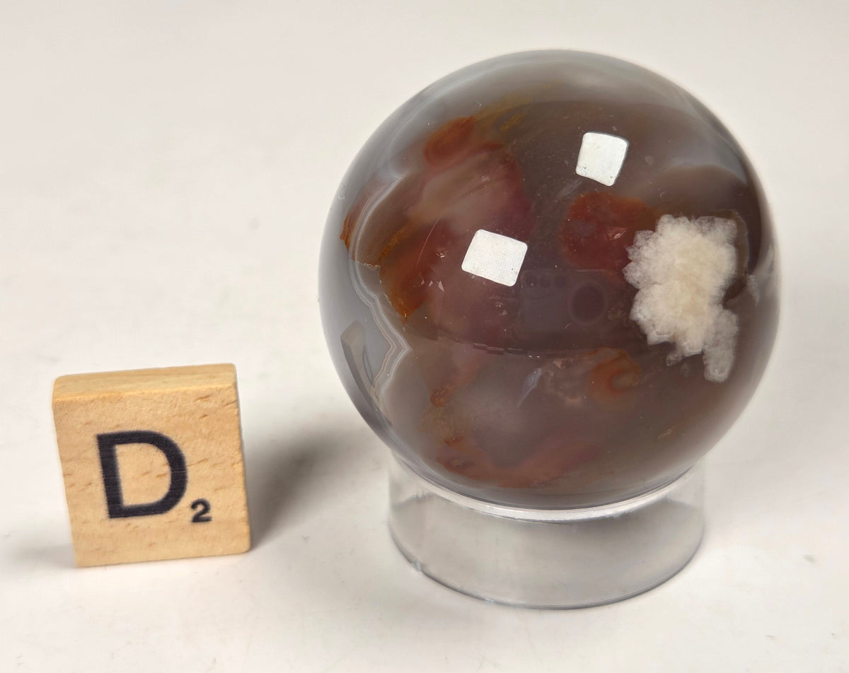 Flower Agate crystal sphere carving with pink and grey coloring, on an acrylic holder, with a scrabble tile against a marbled background.