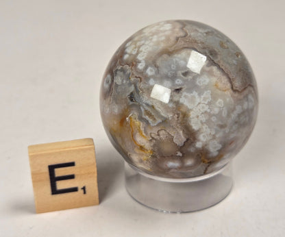 Flower Agate crystal sphere carving with pink and grey coloring, on an acrylic holder, with a scrabble tile against a marbled background.