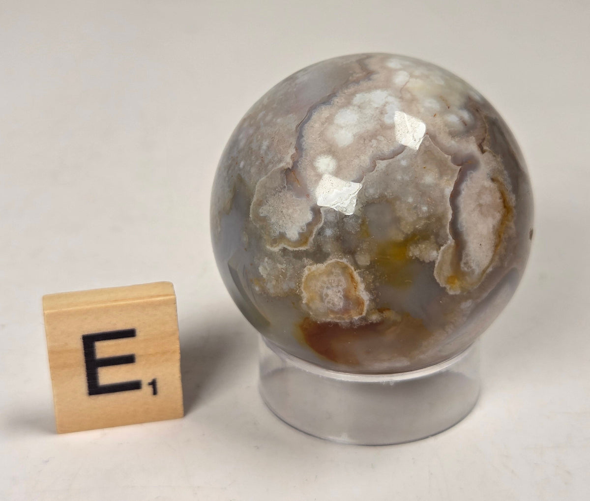 Flower Agate crystal sphere carving with pink and grey coloring, on an acrylic holder, with a scrabble tile against a marbled background.