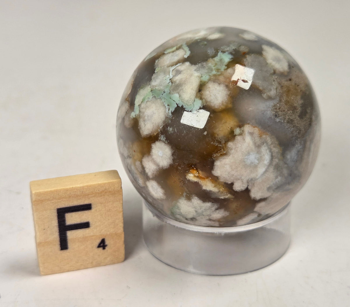Flower Agate crystal sphere carving with pink and grey coloring, on an acrylic holder, with a scrabble tile against a marbled background.
