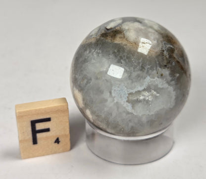 Flower Agate crystal sphere carving with pink and grey coloring, on an acrylic holder, with a scrabble tile against a marbled background.
