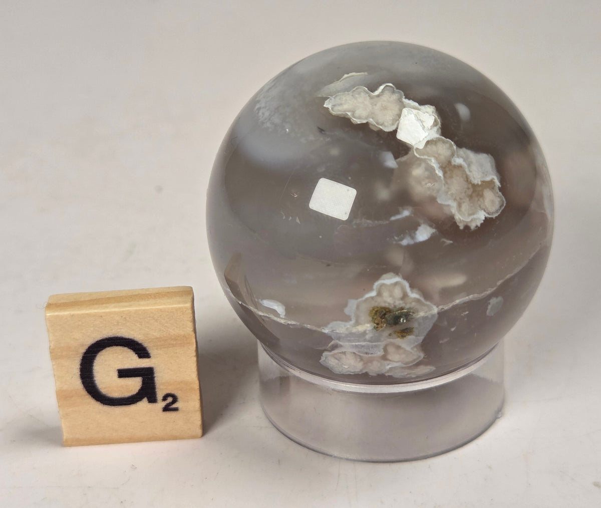 Flower Agate crystal sphere carving with pink and grey coloring, on an acrylic holder, with a scrabble tile against a marbled background.