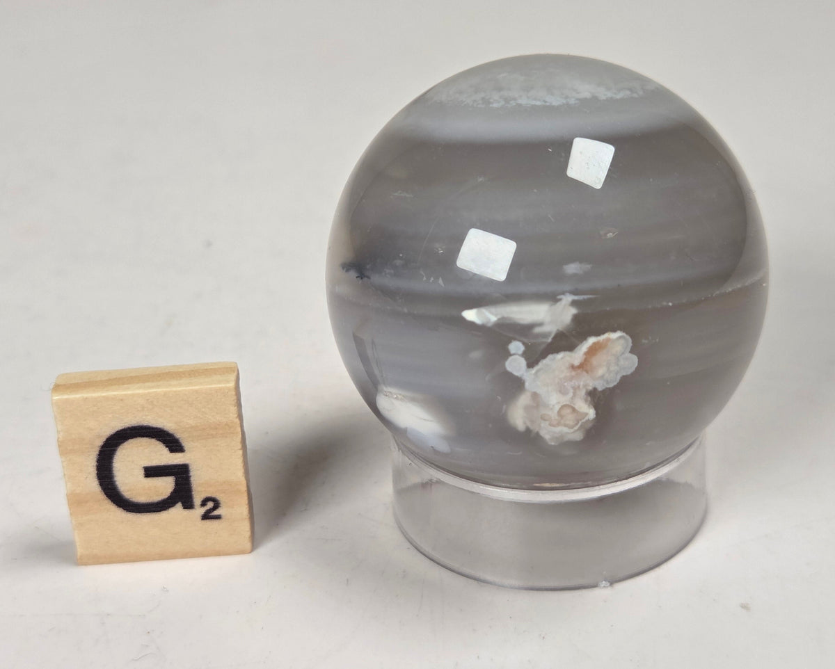 Flower Agate crystal sphere carving with pink and grey coloring, on an acrylic holder, with a scrabble tile against a marbled background.