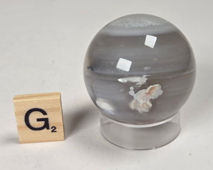 Flower Agate crystal sphere carving with pink and grey coloring, on an acrylic holder, with a scrabble tile against a marbled background.
