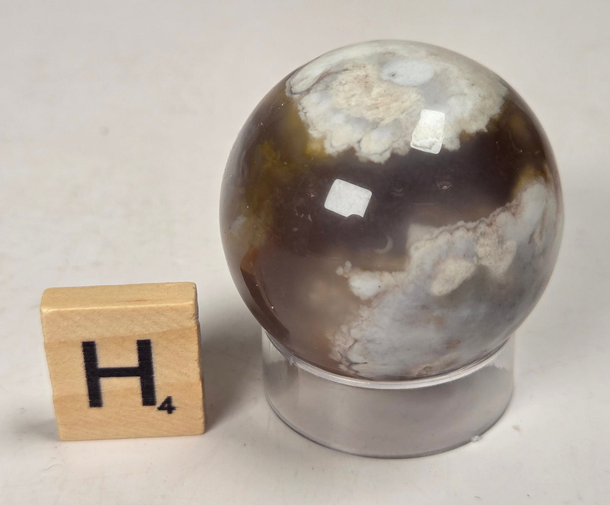 Flower Agate crystal sphere carving with pink and grey coloring, on an acrylic holder, with a scrabble tile against a marbled background.