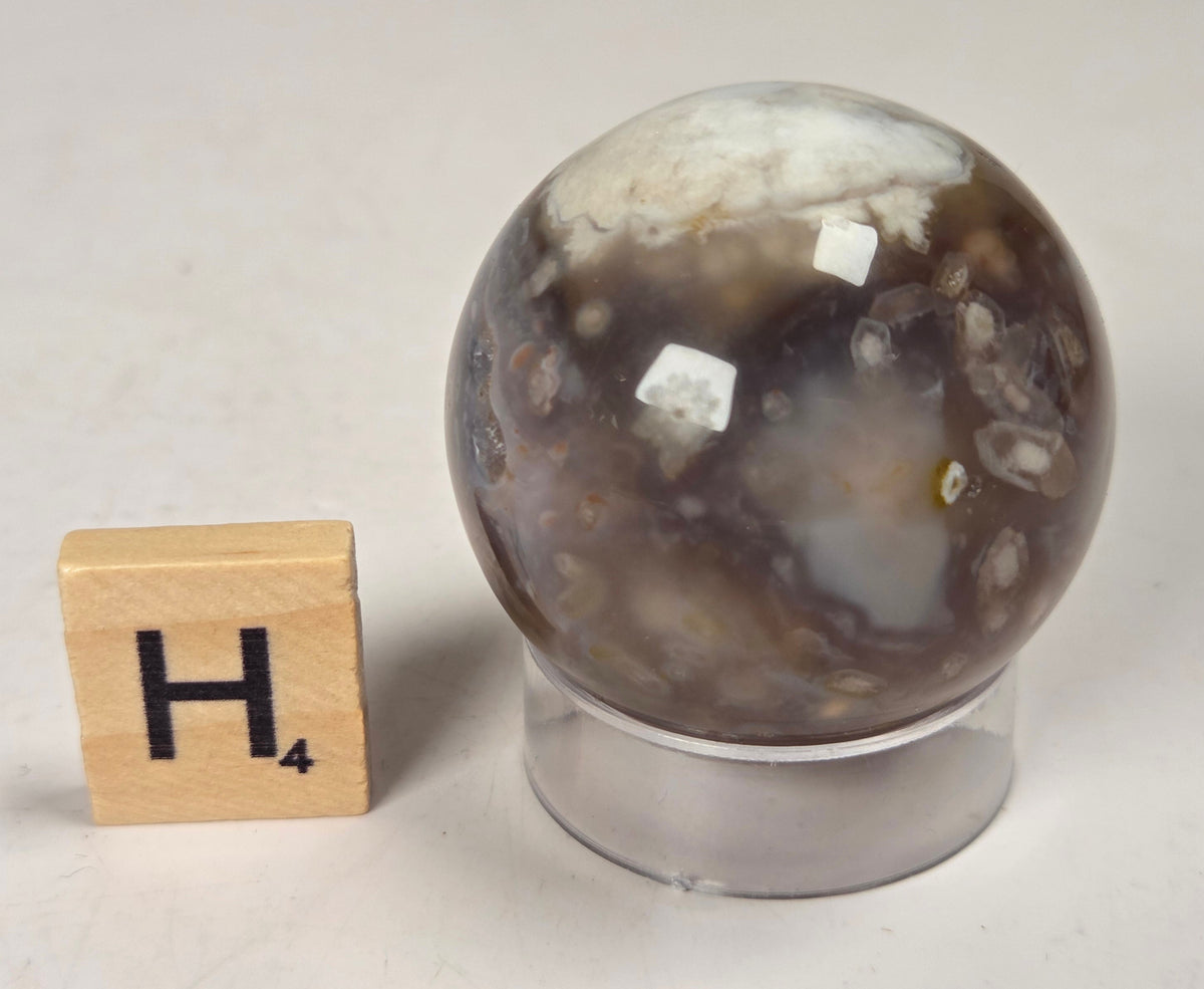 Flower Agate crystal sphere carving with pink and grey coloring, on an acrylic holder, with a scrabble tile against a marbled background.
