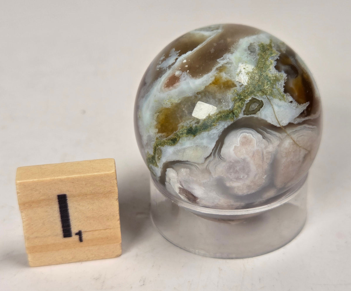 Flower Agate crystal sphere carving with pink and grey coloring, on an acrylic holder, with a scrabble tile against a marbled background.