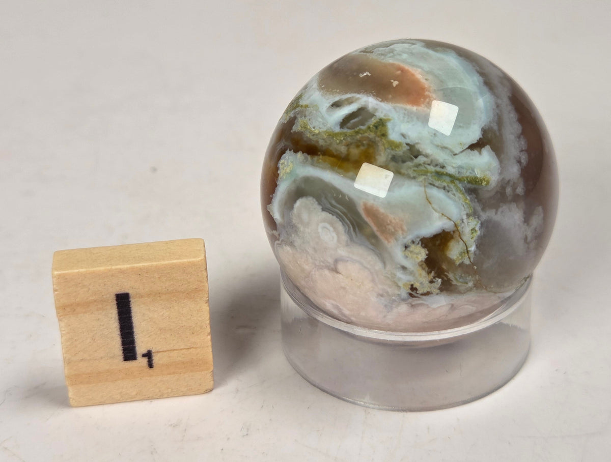 Flower Agate crystal sphere carving with pink and grey coloring, on an acrylic holder, with a scrabble tile against a marbled background.