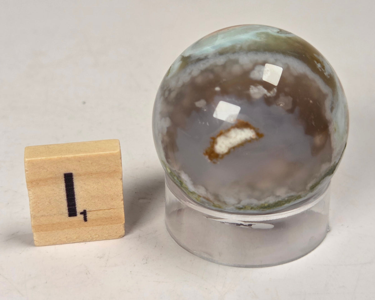 Flower Agate crystal sphere carving with pink and grey coloring, on an acrylic holder, with a scrabble tile against a marbled background.