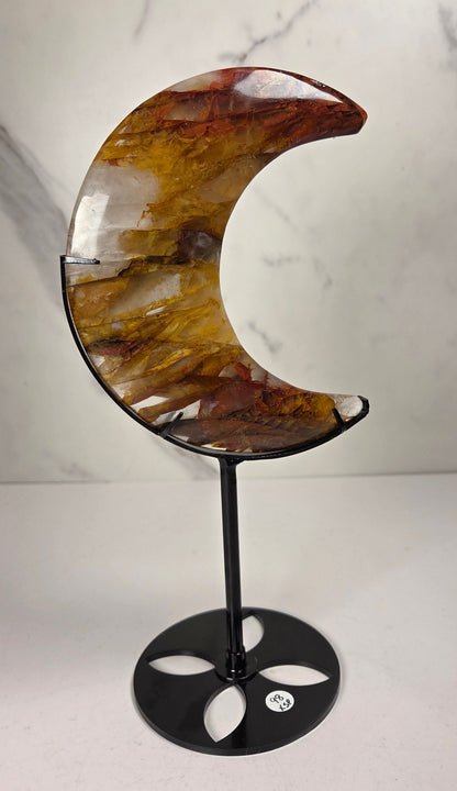 Crystal carved moon with red and yellow inclusions on a black stand against a marbled background