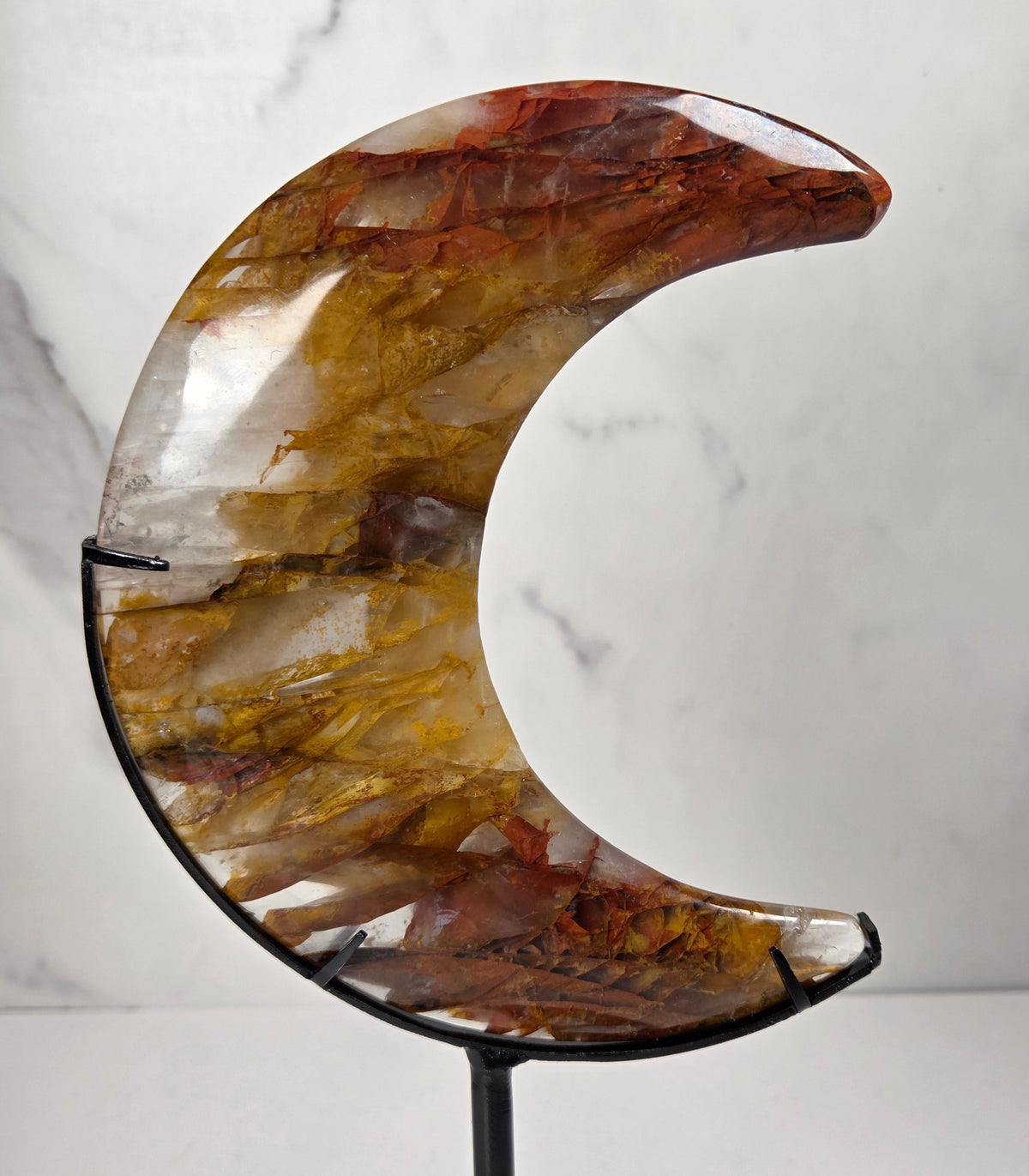 Crystal carved moon with red and yellow inclusions on a black stand against a marbled background