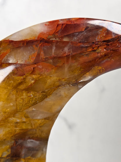 Close-up of a Crystal carved moon with red and yellow inclusions on a black stand against a marbled background