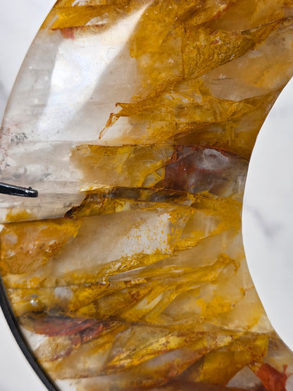 Close-up of a Crystal carved moon with red and yellow inclusions on a black stand against a marbled background