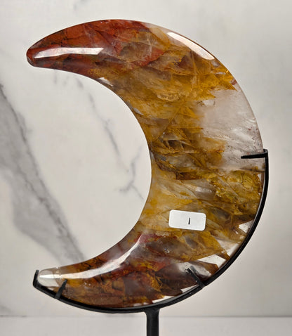 Crystal carved moon with red and yellow inclusions on a black stand against a marbled background