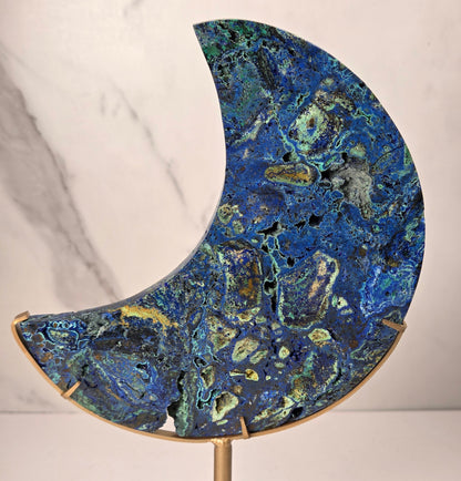 Crystal moon carving with green and blue coloring on a gold stand against a marbled background