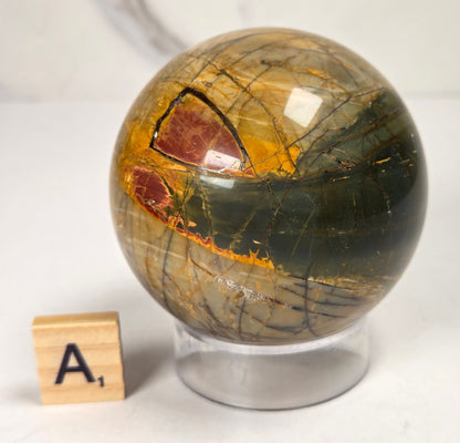 Polished Cherry Creek jasper sphere in various shades of red, orange, yellow, and green, with acrylic holder and a scrabble tile on a white background