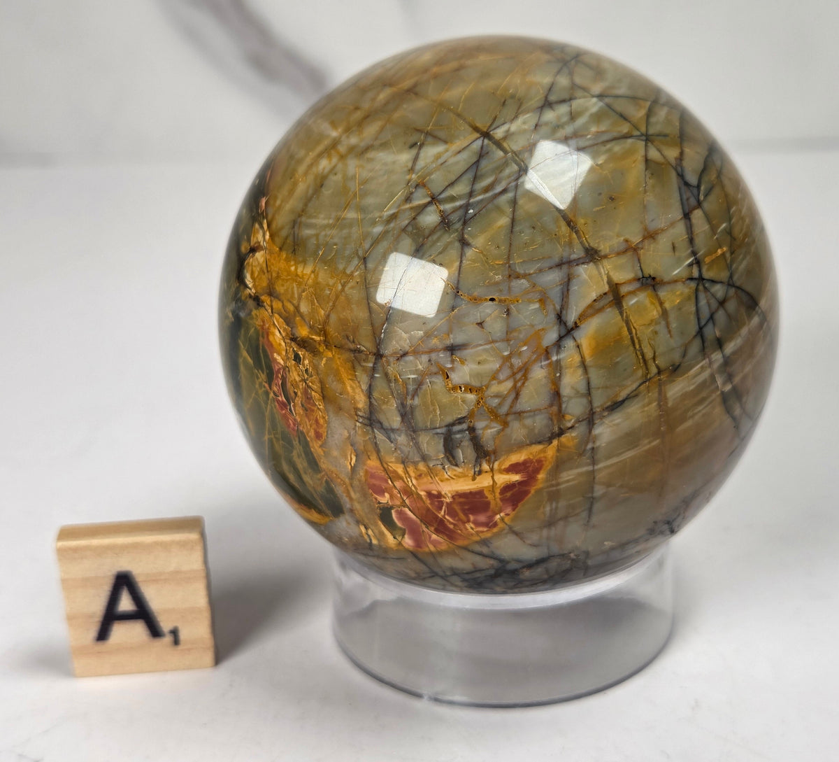 Polished Cherry Creek jasper sphere in various shades of red, orange, yellow, and green, with acrylic holder and a scrabble tile on a white background