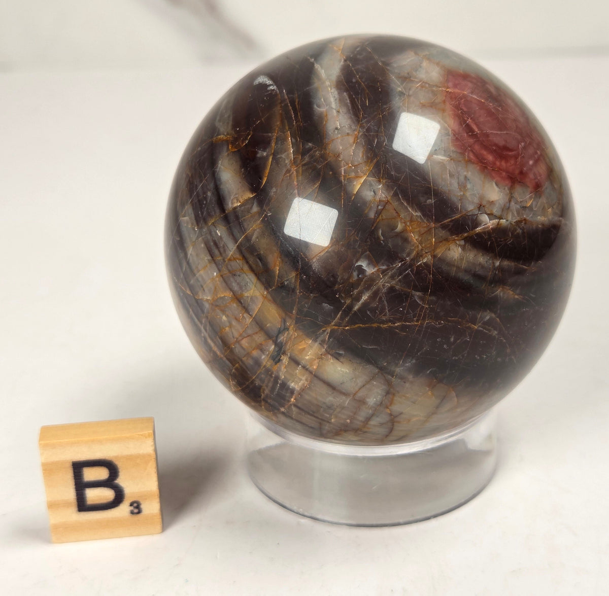 Polished Cherry Creek jasper sphere in various shades of red, orange, yellow, and green, with acrylic holder and a scrabble tile on a white background