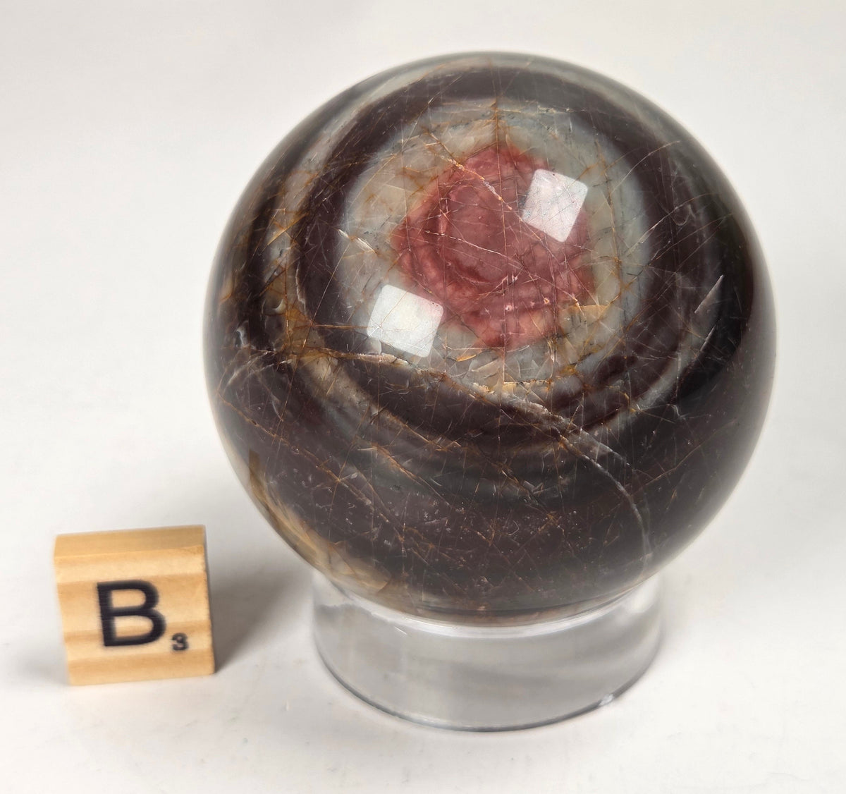 Polished Cherry Creek jasper sphere in various shades of red, orange, yellow, and green, with acrylic holder and a scrabble tile on a white background